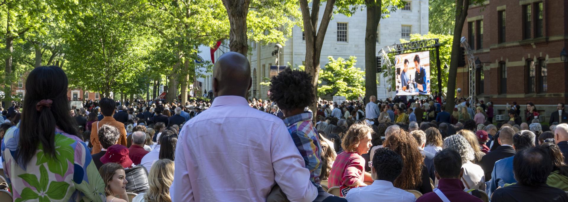 Families gather at Commencement with a video screen in the background.